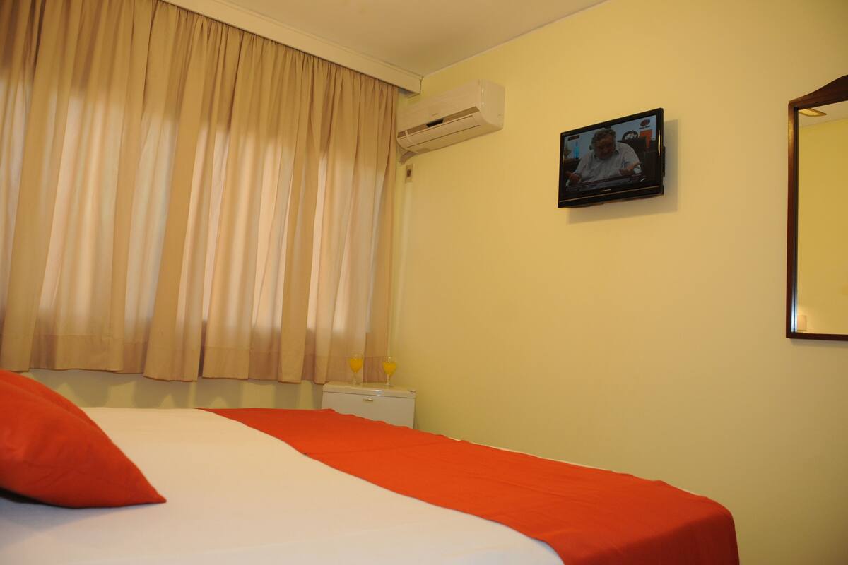 A hotel room with a bed, curtains, an air conditioner, a television, and a small table with a bottle and glasses.