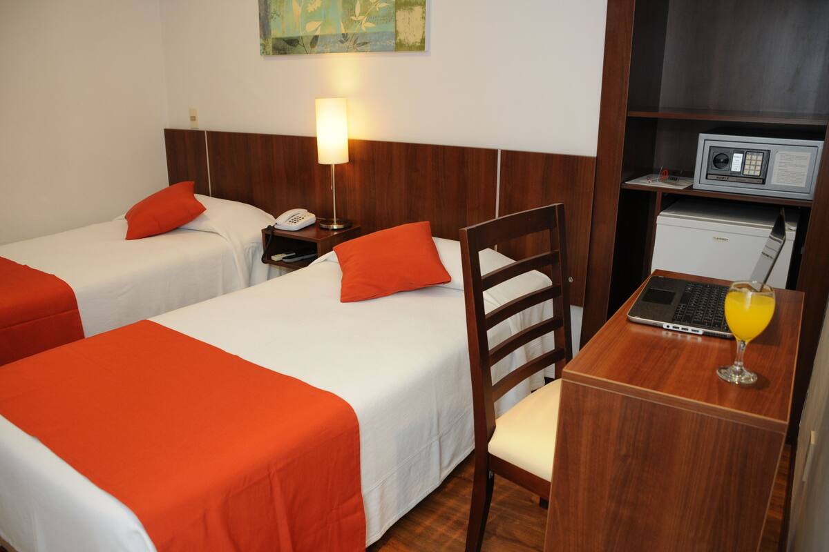 A hotel room with two beds, each with an orange bedspread, a bedside lamp, a desk with a laptop and a glass of orange juice, and a small refrigerator.