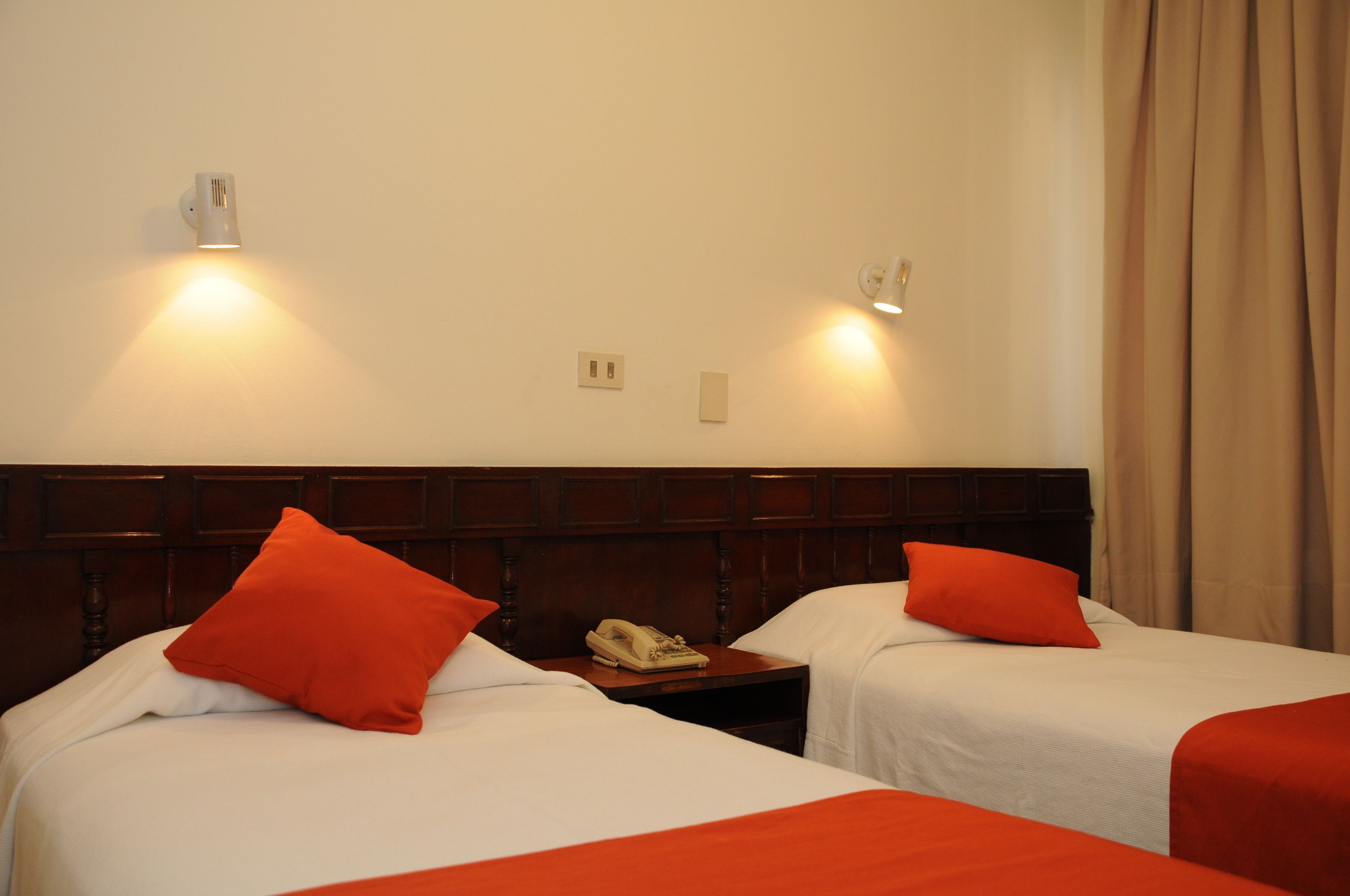 A hotel room with two beds, each with a red pillow, a wooden headboard, and a bedside table with a telephone.