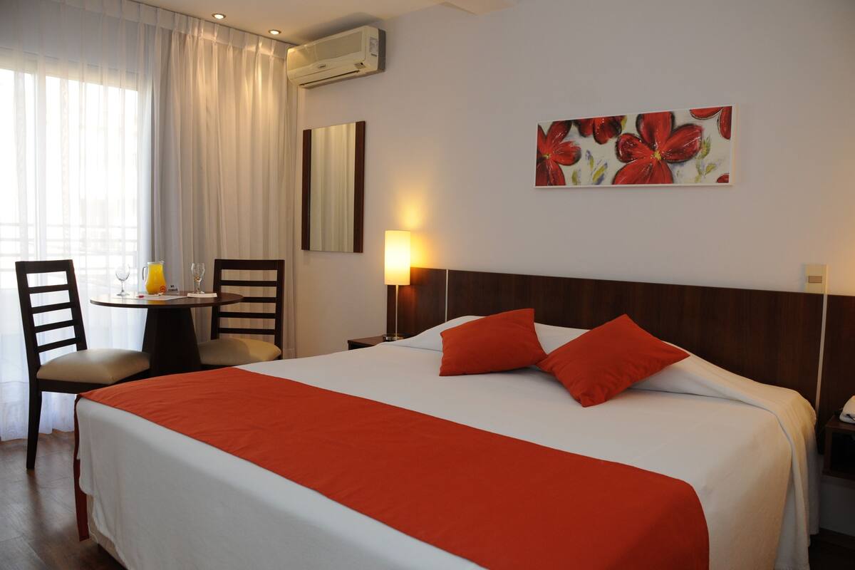 A hotel room with a large bed, two red pillows, a wooden headboard, a bedside table with a lamp, a chair, and a window with curtains.