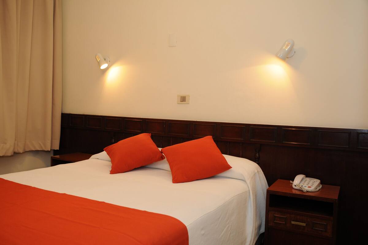 A hotel room with a large bed, two red pillows, a wooden headboard, a bedside table with a telephone, and wall-mounted lights.