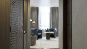 Una habitación de hotel moderna con piso de madera tipo espiga, una zona de estar con dos sillones y una mesita, y un ventanal con cortinas.