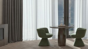 Una habitación moderna con una mesa redonda de madera, dos sillas verdes, un ventanal con cortinas translúcidas y vistas al exterior.