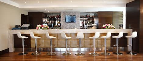 A bar area with a marble countertop, six white bar stools, a television mounted on the wall, and a variety of bottles and glasses on shelves.