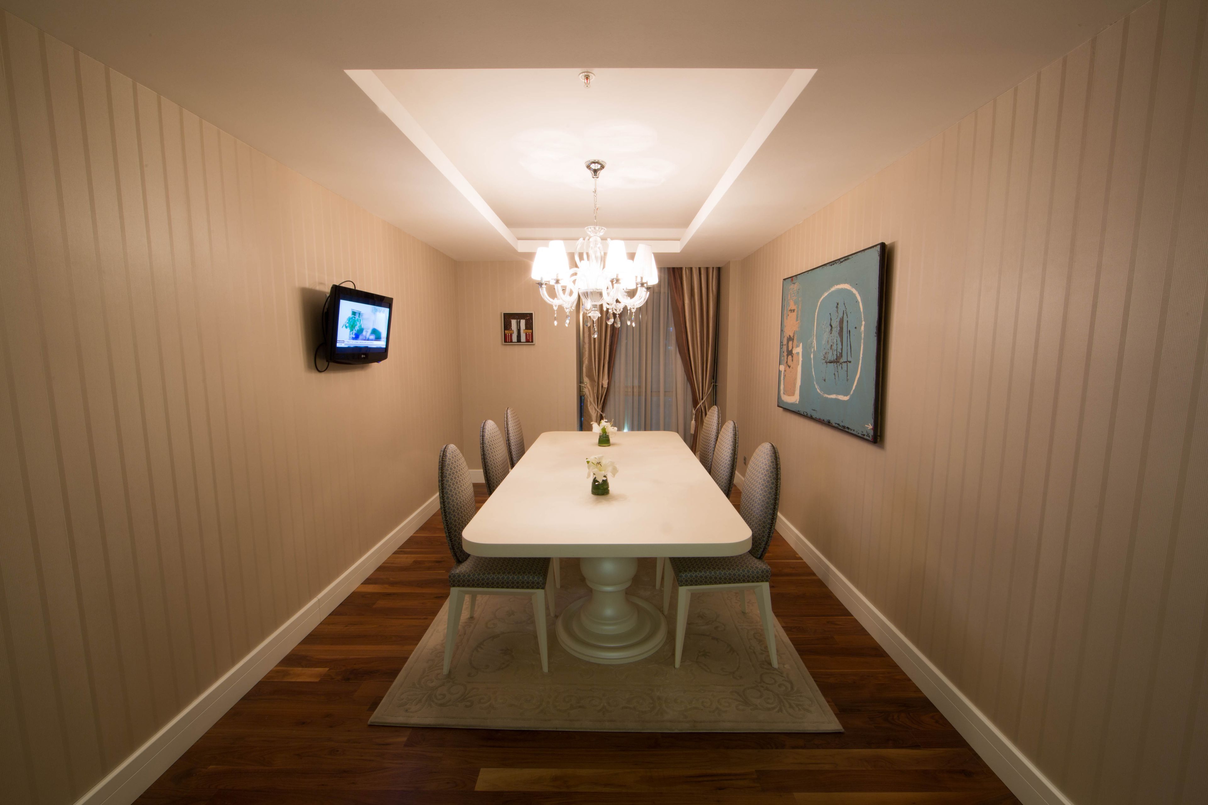 A dining room with a rectangular table, six chairs, a chandelier, a flat-screen TV, and a framed artwork on the wall.
