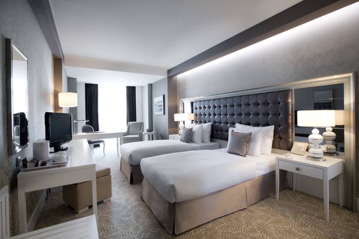 Ein Hotelzimmer mit einem großen Bett, einem kleineren Bett, einem Schreibtisch mit Computer, einem Fernseher und einem Spiegel.