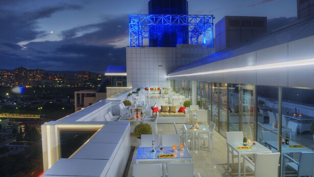 Un bar situé sur un toit, avec une terrasse, une vue sur la ville et une tour dont l’architecture distinctive est illuminée de lumière bleue.