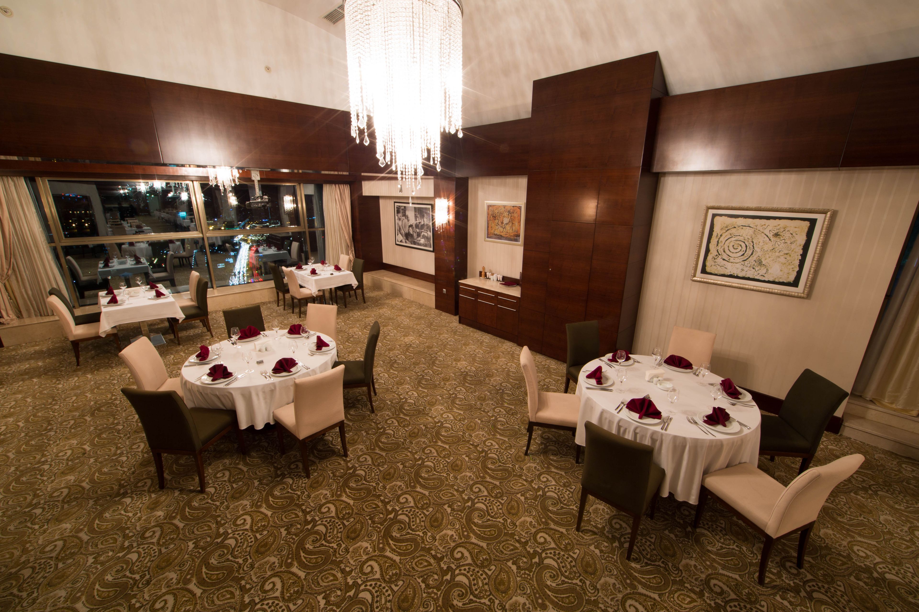 A dining area with round tables set for four, each with white tablecloths and red napkins. The room has a patterned carpet and large windows offering a view of the cityscape.