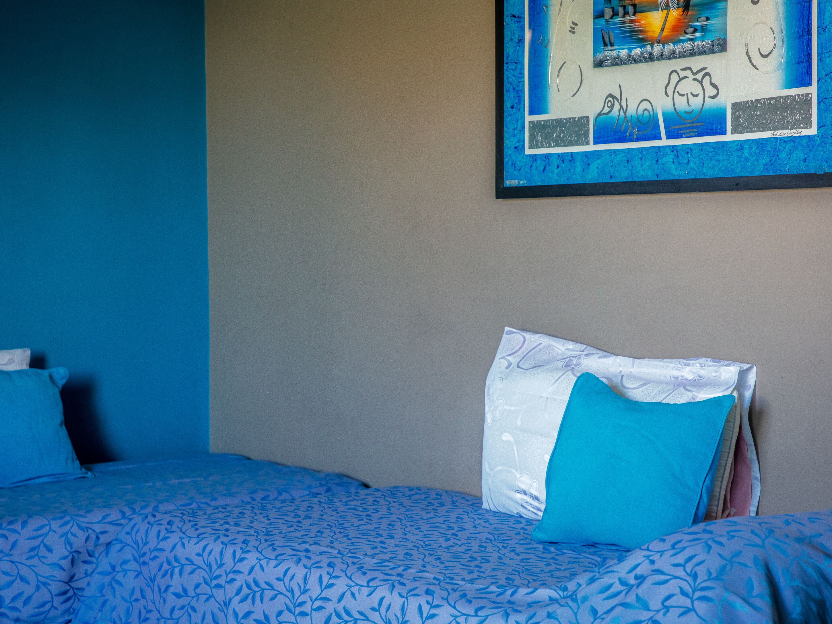 Habitación de hotel con una cama que tiene una colcha y almohadas de diseño azul, una pared con una obra de arte enmarcada, y una pared lisa al fondo.