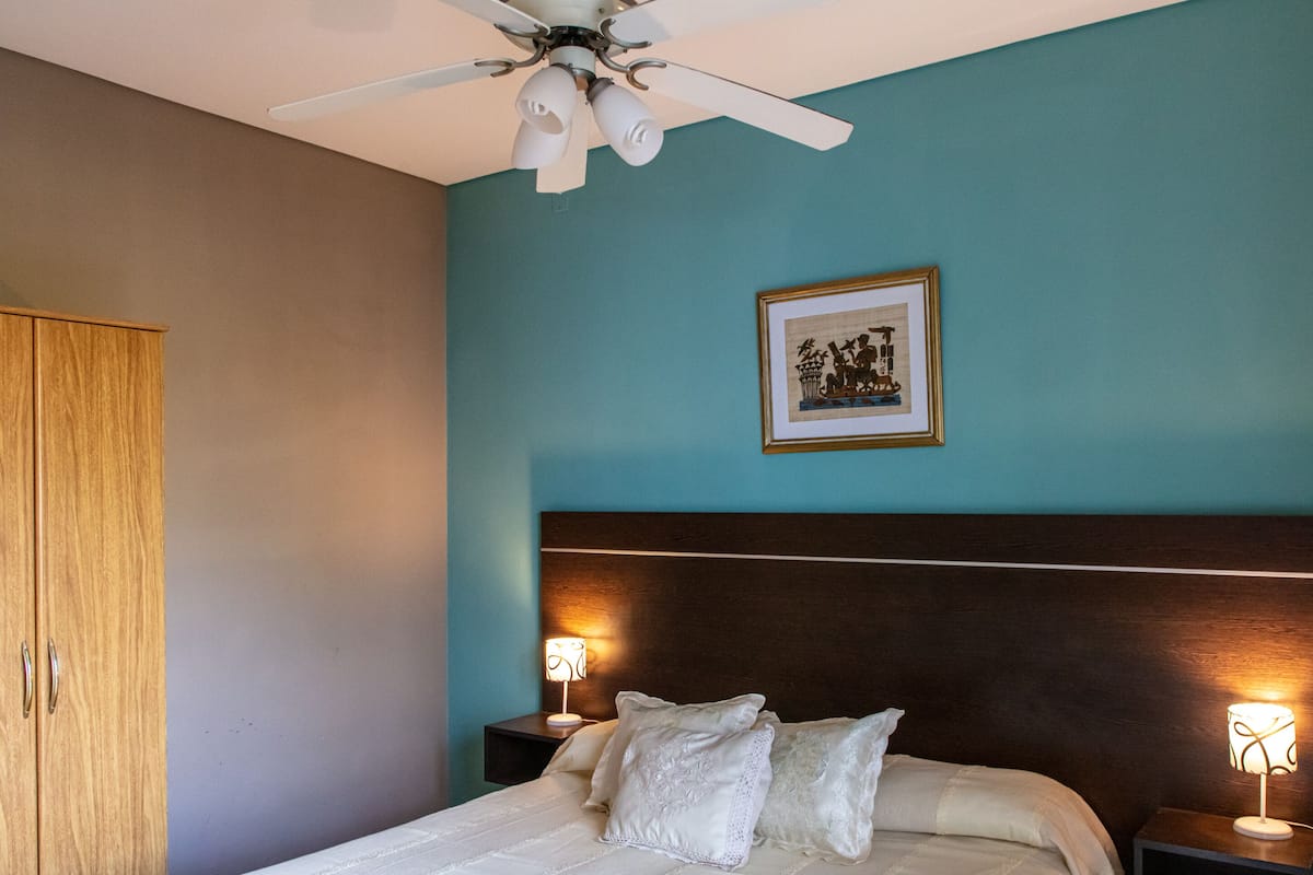 Ein Schlafzimmer mit einem Bett, einem Deckenventilator, einem gerahmten Bild an der Wand und zwei Nachttischlampen.