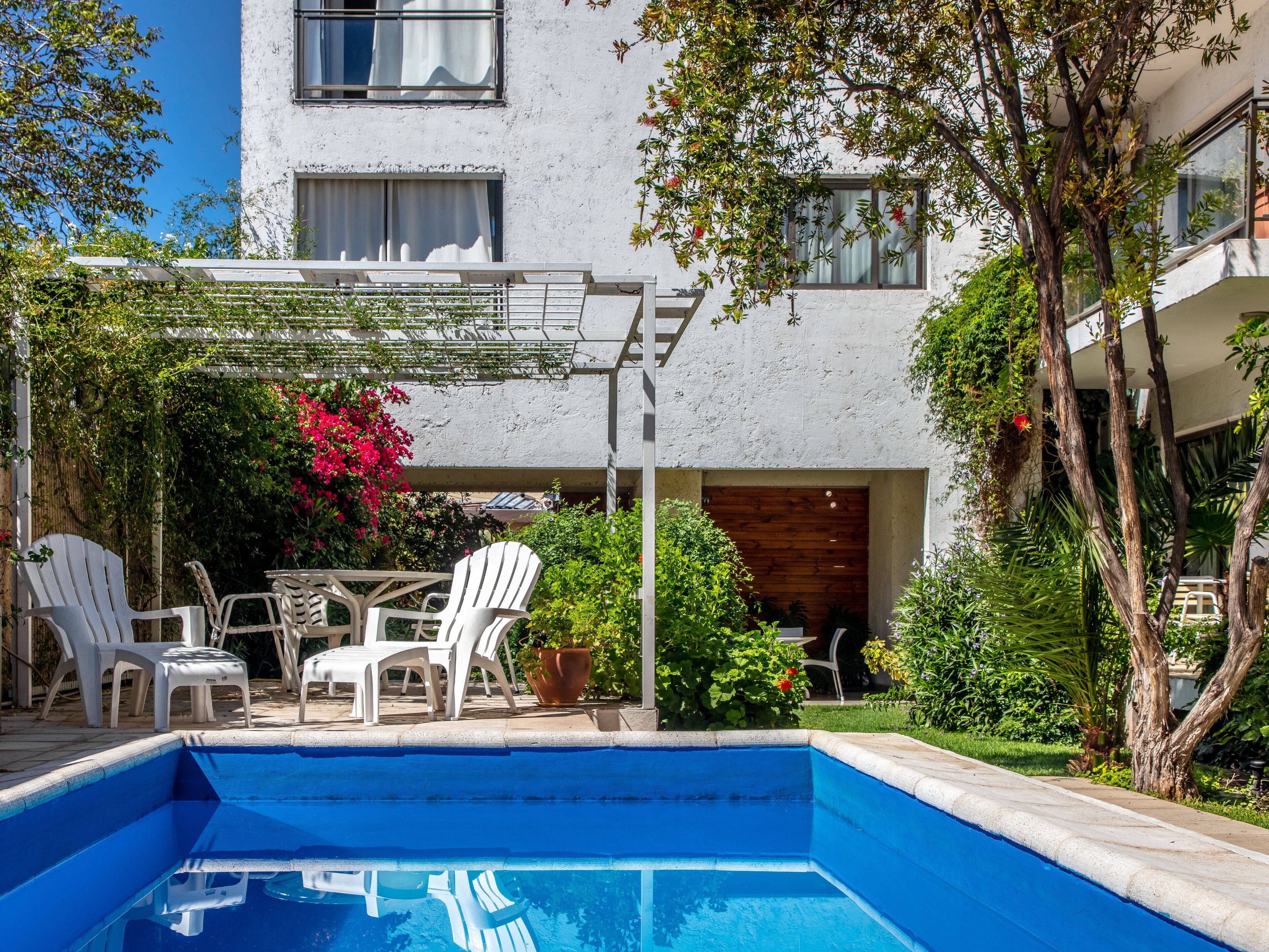 Ein Swimmingpool mit weißen Stühlen und einem Tisch, umgeben von Grünflächen und einem Gebäude mit Fenstern.