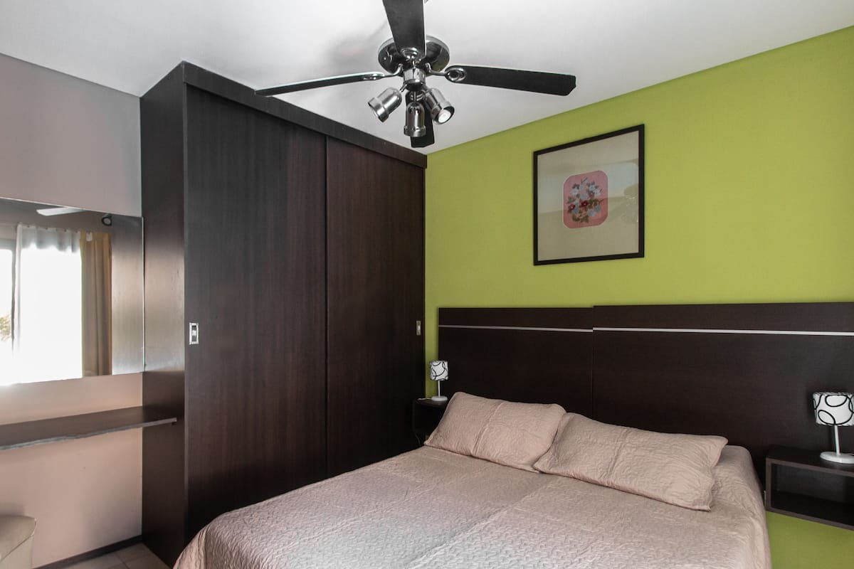 Ein Schlafzimmer mit Bett, Deckenventilator, Nachttisch, gerahmtem Bild an der Wand und Kleiderschrank.