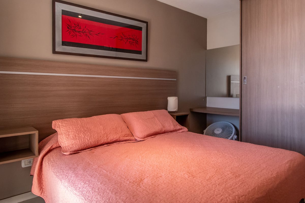 Ein ordentlich bezogenes Bett mit einer rosa Tagesdecke, ein gerahmtes Kunstwerk darüber und eine Holzgarderobe zur Rechten.