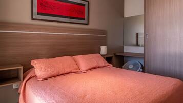 Un lit bien fait, recouvert d’une couvre-lit rose, avec une œuvre d’art encadrée au-dessus, et une armoire en bois sur le côté droit.