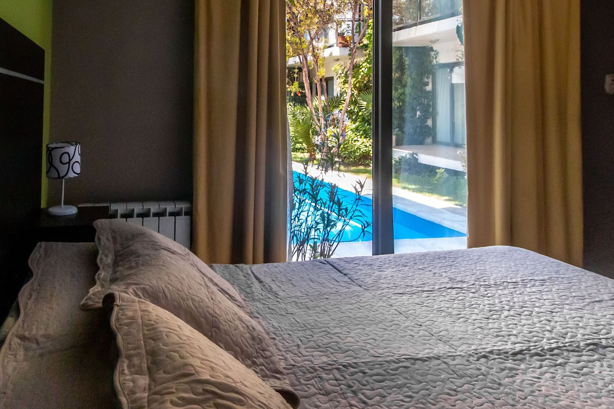 Ein Bett mit weißer Strukturdecke, ein Nachttisch mit Lampe und Blick auf einen Swimmingpool durch ein Fenster mit Vorhängen.