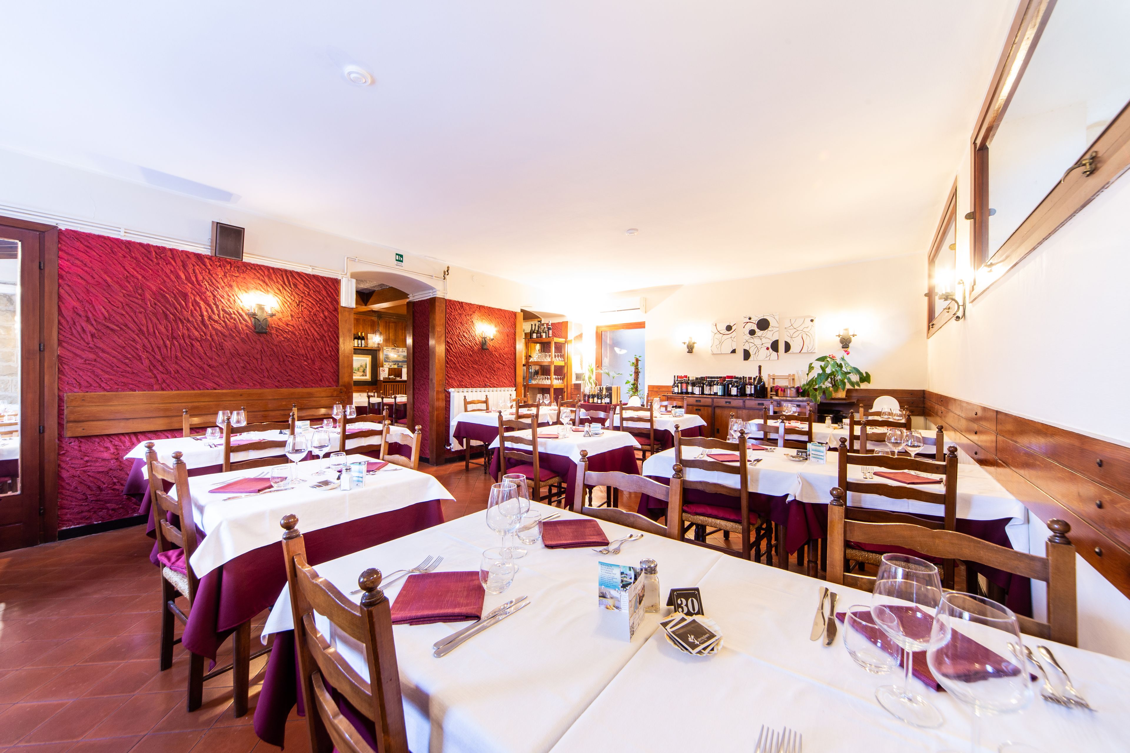 A restaurant with multiple tables set for dining, wooden chairs, and a red textured wall.