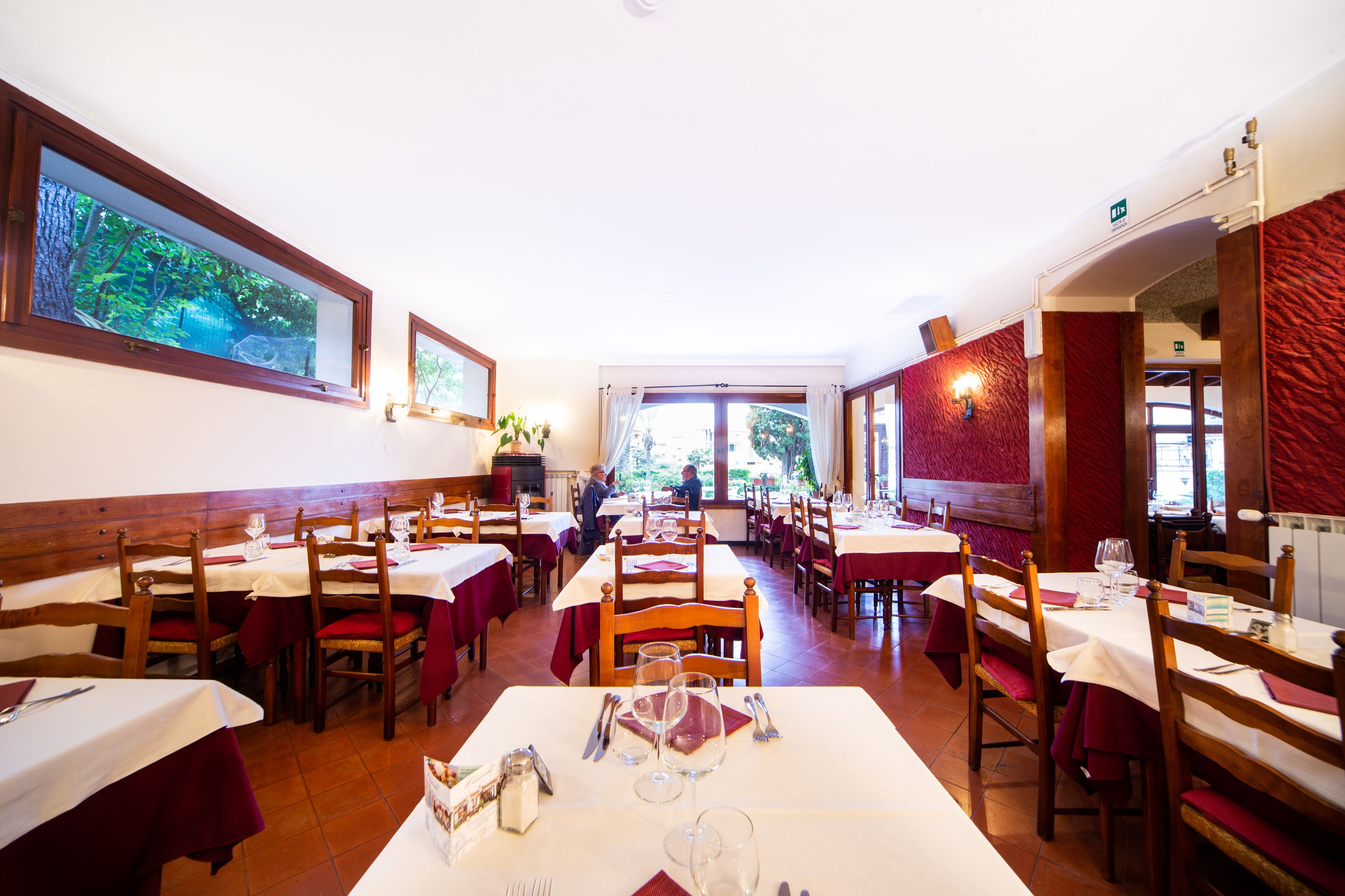 A restaurant with multiple tables and chairs, a large window with a view of greenery, and a red textured wall.