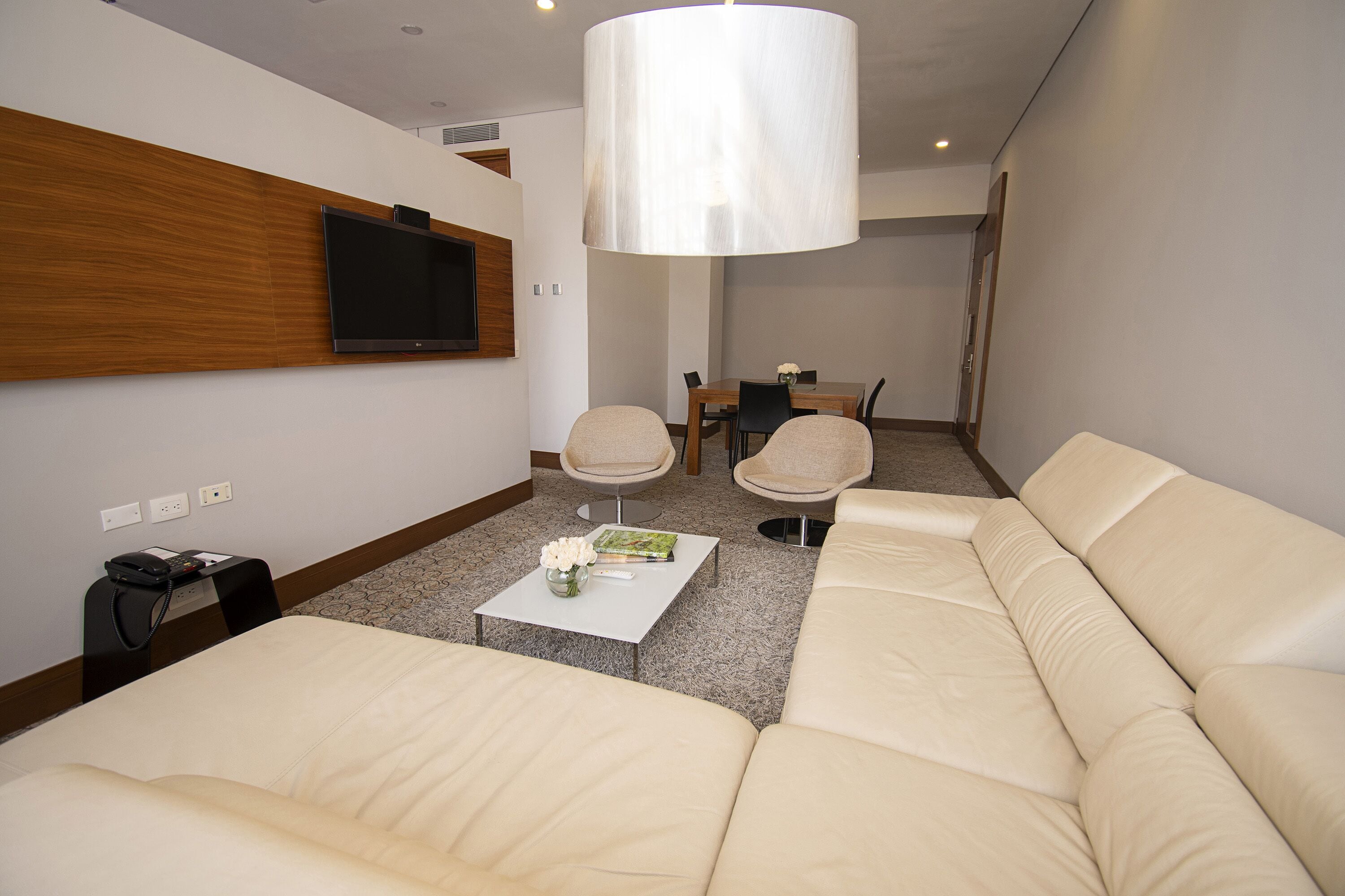 A modern living room with a sofa, a coffee table with a vase and book, a TV mounted on the wall, and a dining area in the background.