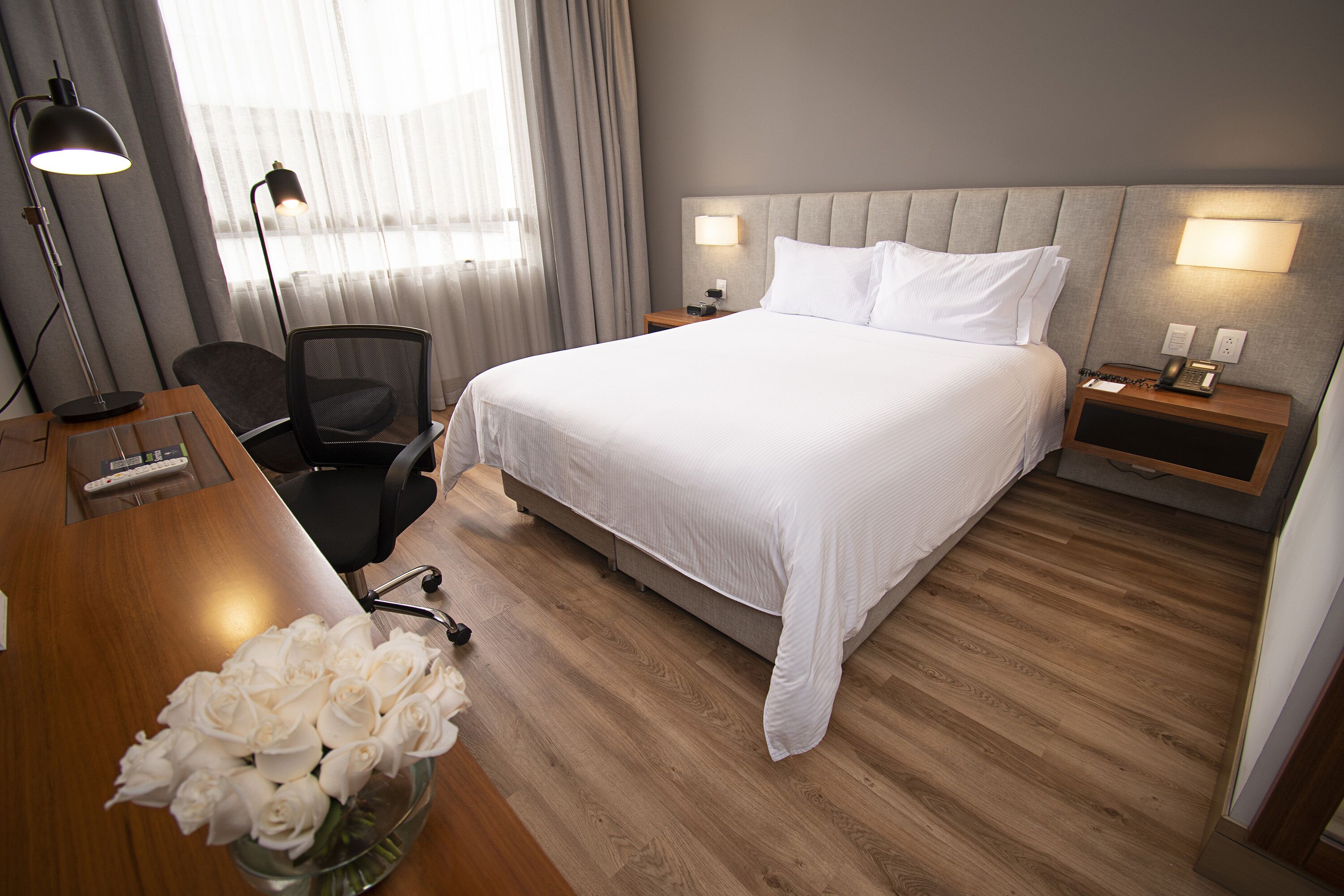 Hotellihuone, jossa on suuri sänky, yöpöydällä olevat lamput, työpöytä tuolilla sekä maljakko valkoisia kukkia.