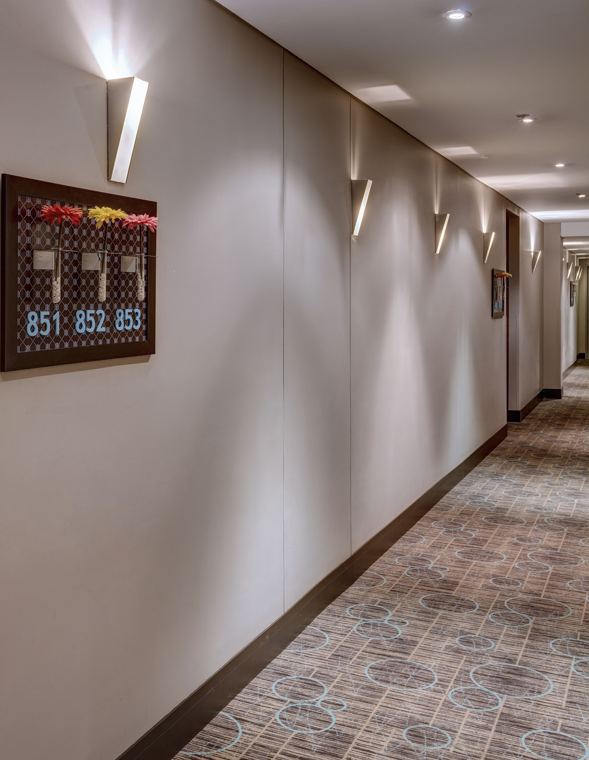 A hotel hallway with a patterned carpet, wall-mounted light fixtures, and a number display board.