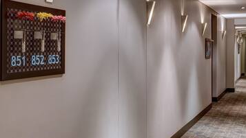 A hotel hallway with a patterned carpet, wall-mounted light fixtures, and a number display board.