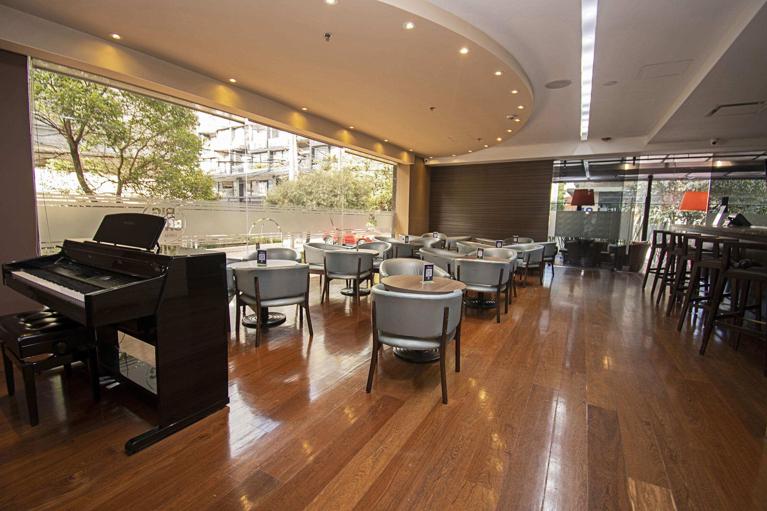 A spacious room with a piano, multiple tables and chairs, and large windows offering a view of the outside.
