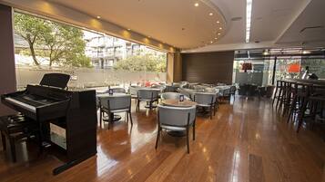 A spacious room with a piano, multiple tables and chairs, and large windows offering a view of the outside.