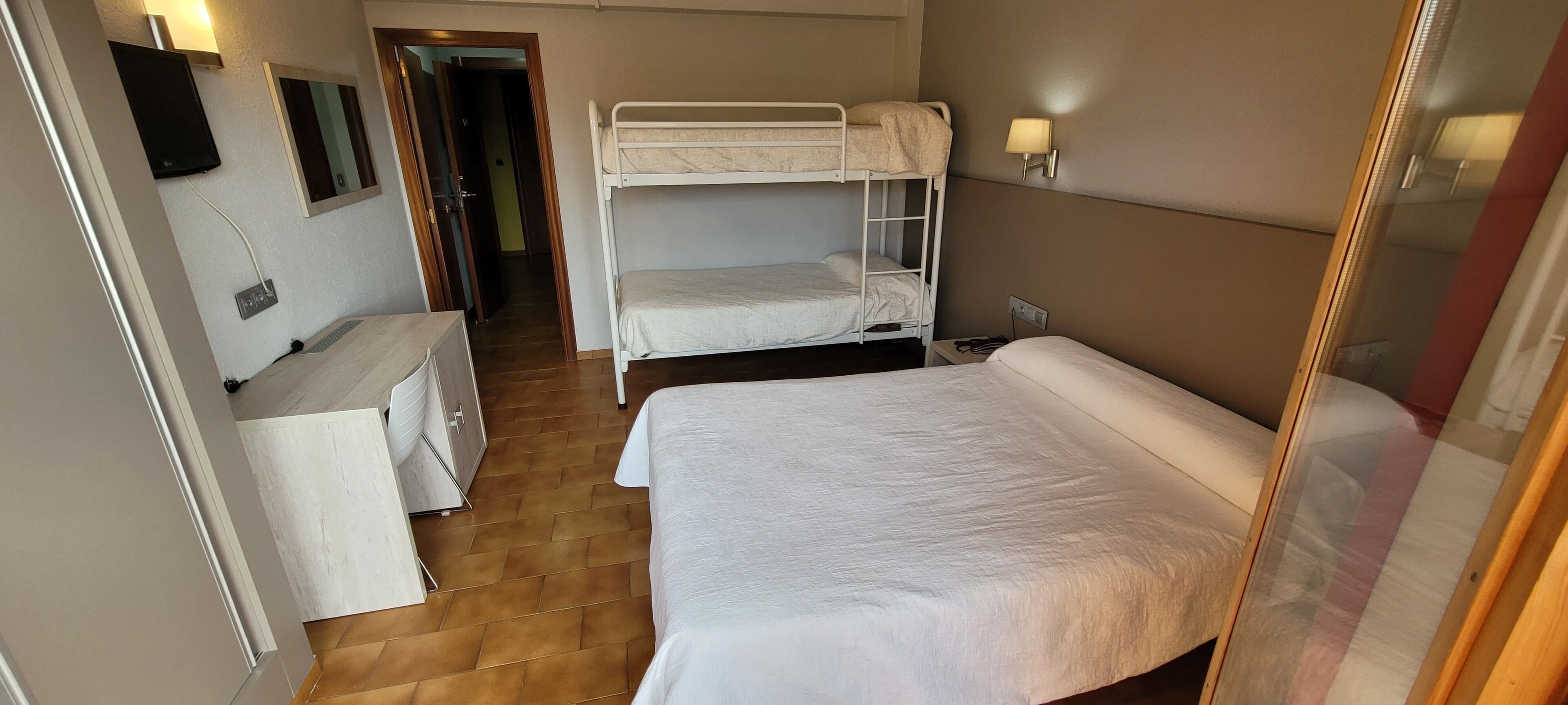 Una habitación de hotel con literas, una cama individual, una mesita de noche, un espejo y un televisor instalado en la pared.