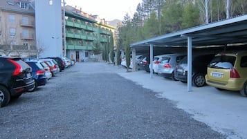Een parkeerplaats met meerdere auto's onder een overkapping, een gebouw met balkons en bomen op de achtergrond.