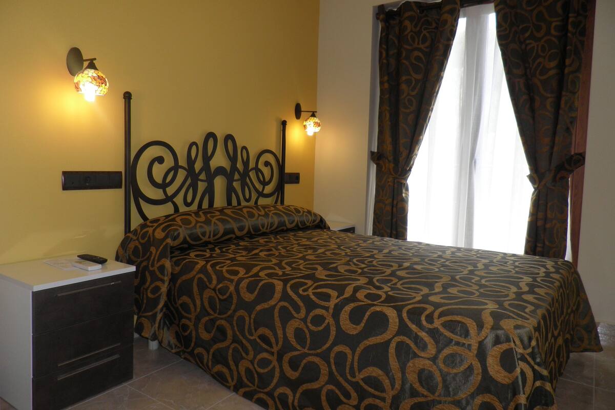 A bedroom with a patterned bedspread, two bedside lamps, a nightstand, and a window with curtains.