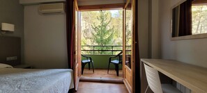 A room with a bed, a desk, a balcony with chairs, and a view of trees.