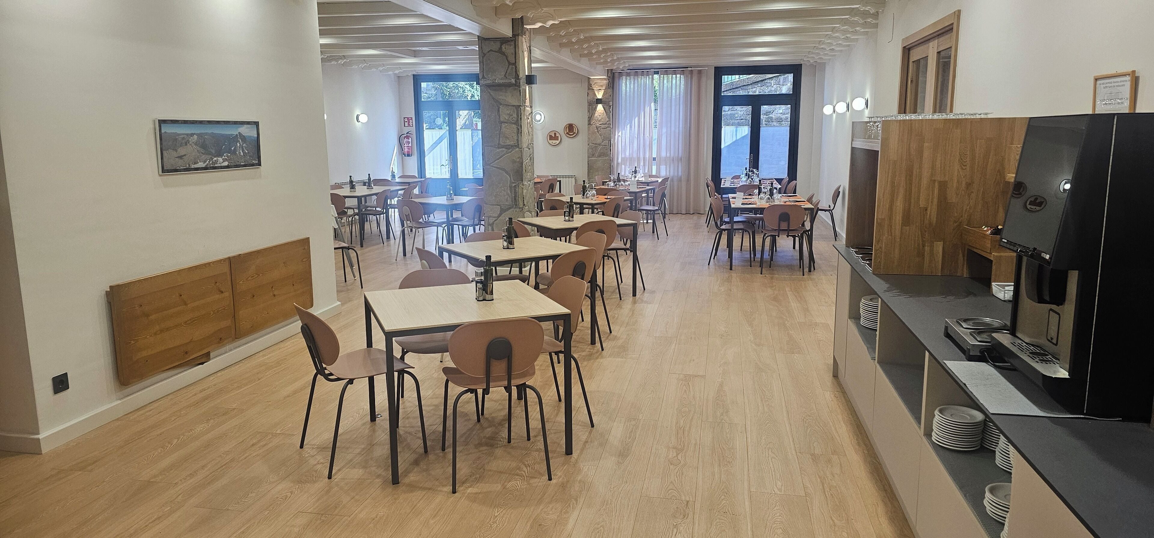 Un comedor moderno con varias mesas y sillas, una encimera de cocina con electrodomésticos, y un ventanal que proporciona luz natural.