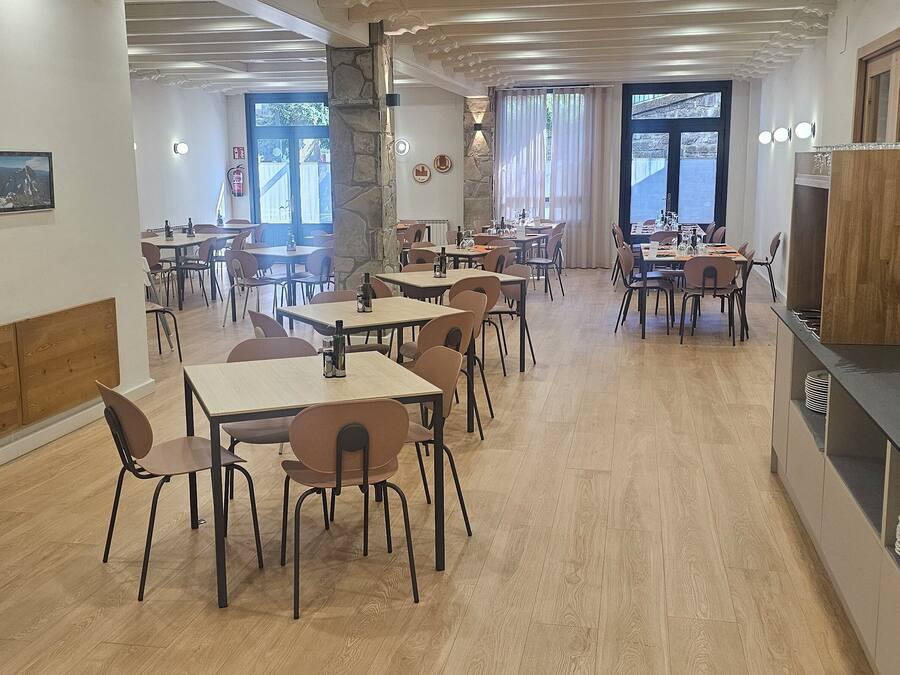 Un comedor moderno con varias mesas y sillas, una encimera de cocina con electrodomésticos, y un ventanal que proporciona luz natural.