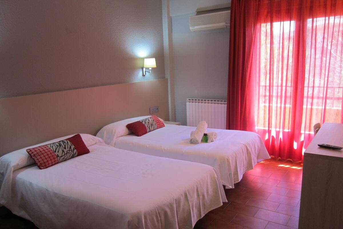 A hotel room with two beds, a desk, a lamp, and a window with red curtains.