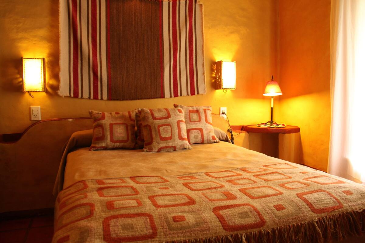 A neatly made bed with patterned pillows and a blanket, a striped wall hanging, and two wall-mounted lamps.