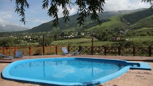 A swimming pool with two lounge chairs, surrounded by a brick patio and a wooden fence, overlooking a valley with houses and mountains. - Posada La Guadalupe (Tafi del Valle)
