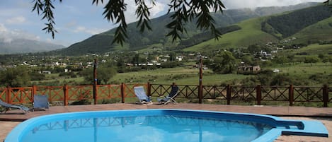 A swimming pool with two lounge chairs, surrounded by a brick patio and a wooden fence, overlooking a valley with houses and mountains.