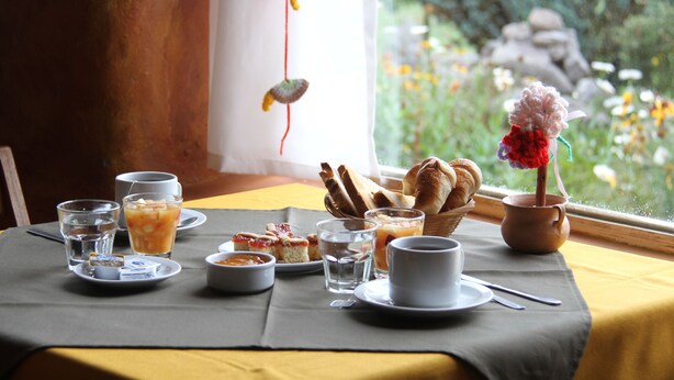 A breakfast setting with a table set for two, including plates, cups, and a basket of bread. - Posada La Guadalupe (Tafi del Valle)