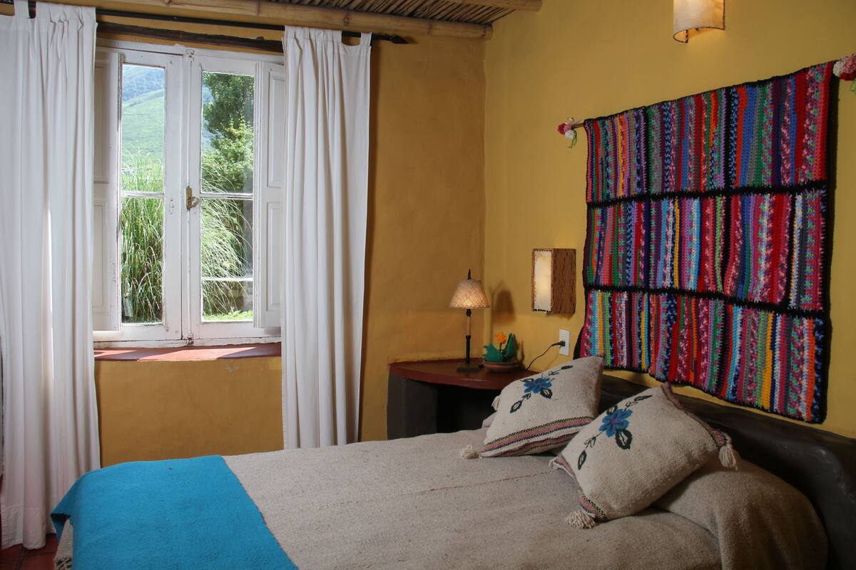 A bedroom with a bed, a window with curtains, a lamp, and a colorful tapestry on the wall.