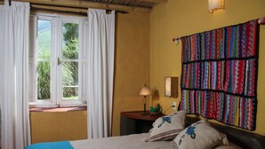 A room with a bed, a window with curtains, a lamp, and a colorful quilt on the wall.