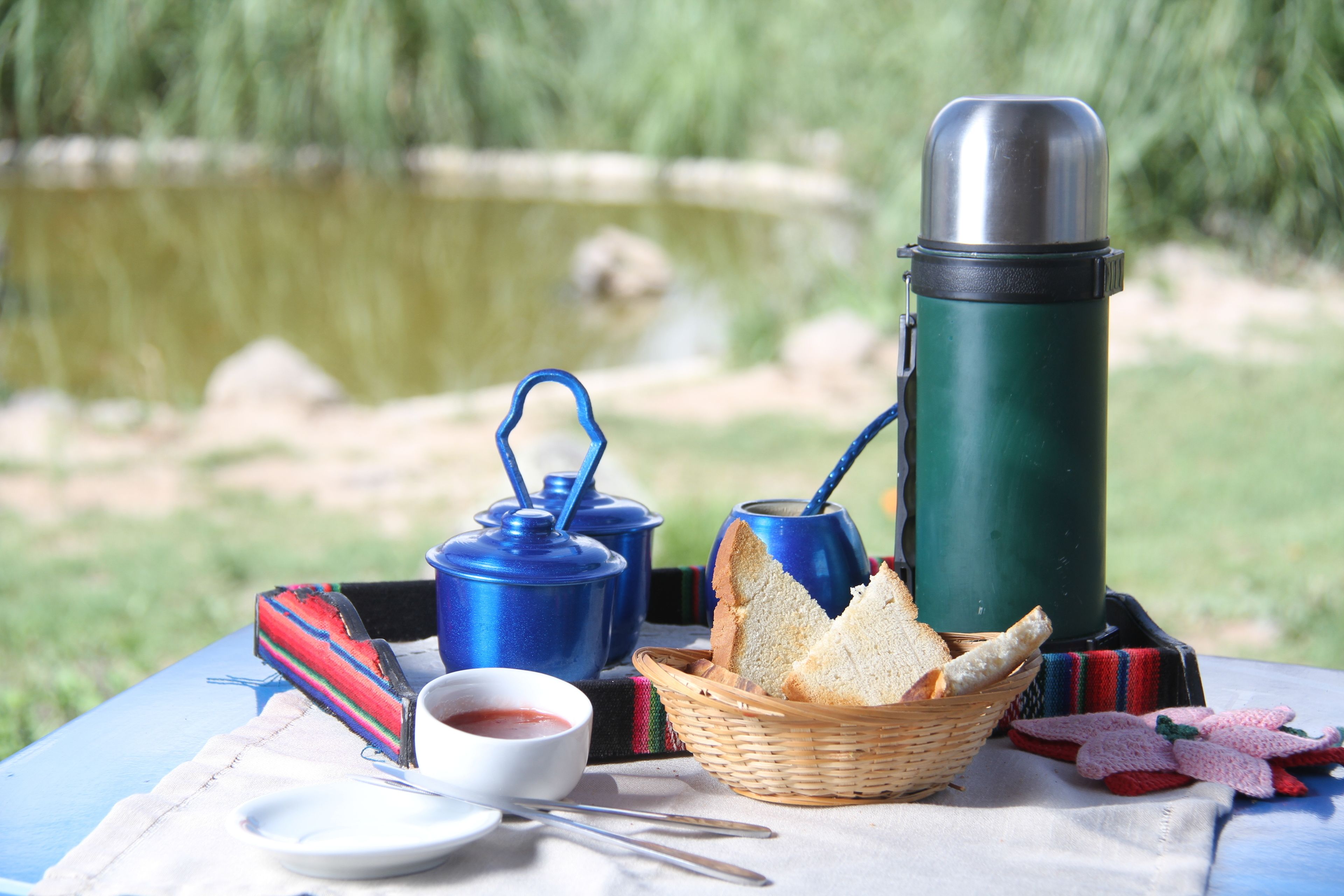 Un montaje para picnic con un termo, tetera, pan y un tazón pequeño sobre una mesa al aire libre.