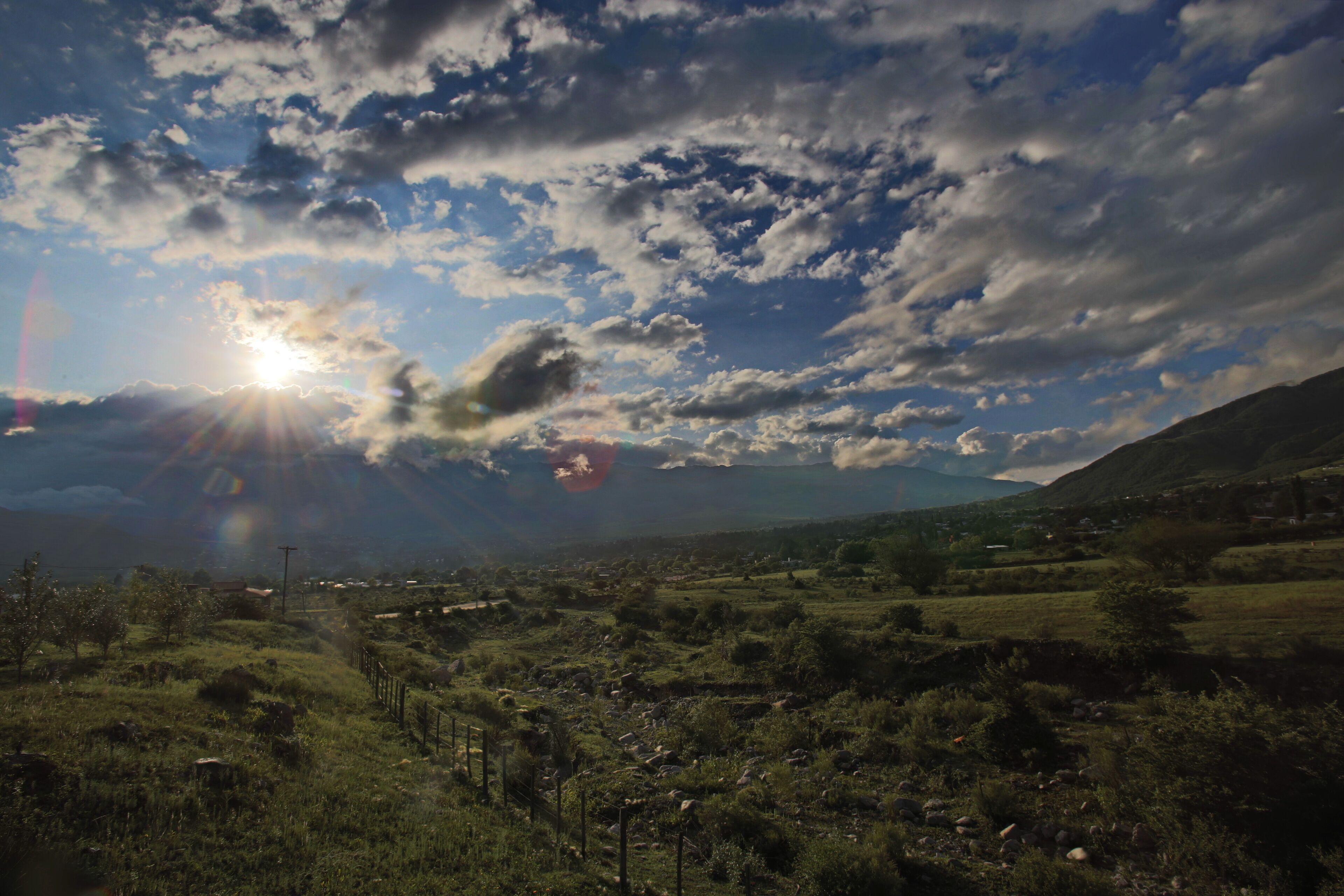 A rural landscape with a clear sky, scattered clouds, and a sun shining through.