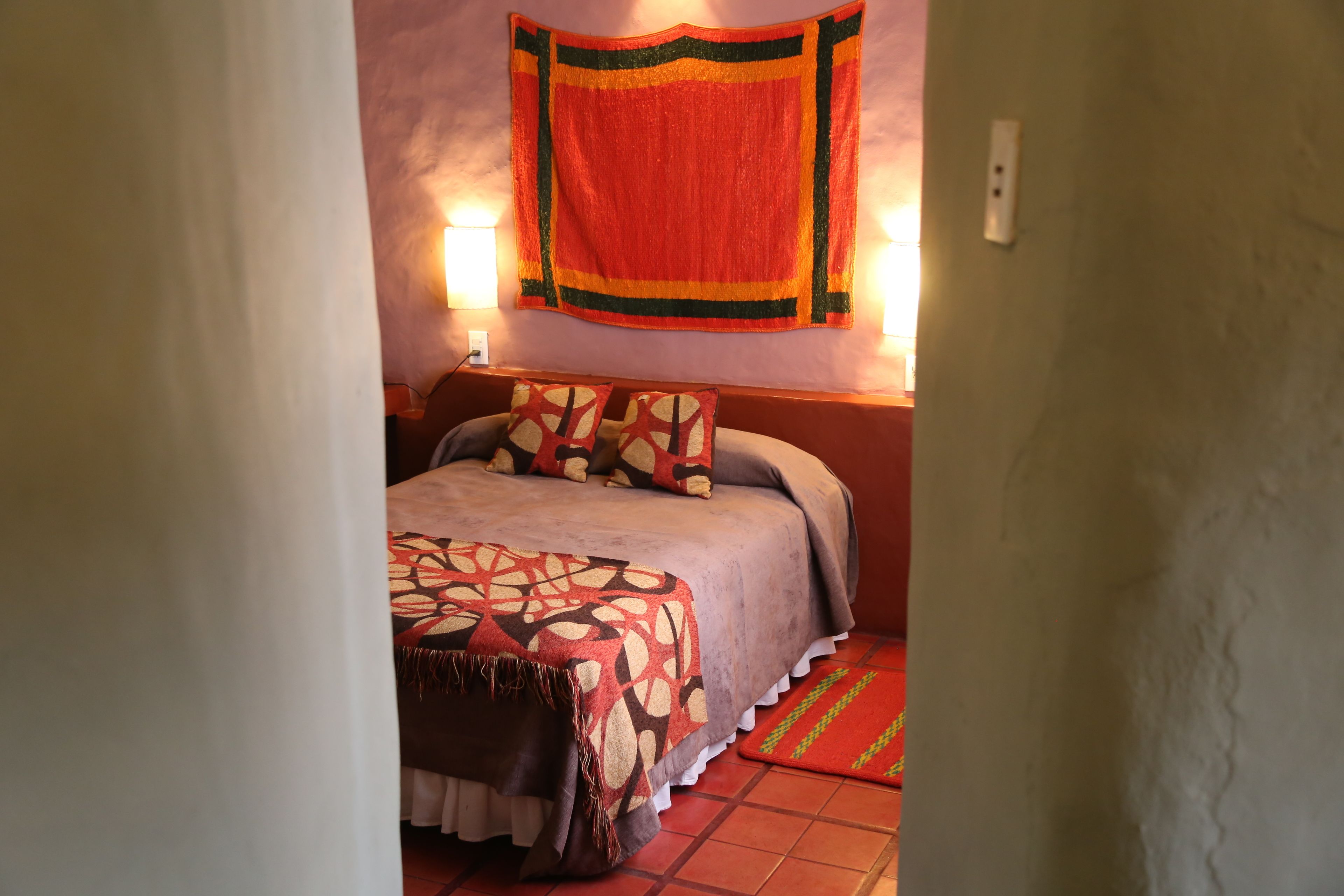 A bedroom with a bed, pillows, a blanket, a wall hanging, and two wall-mounted lamps.