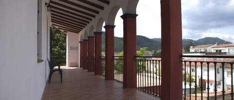 A covered balcony with red pillars and a tiled floor, overlooking a residential area and hills.
