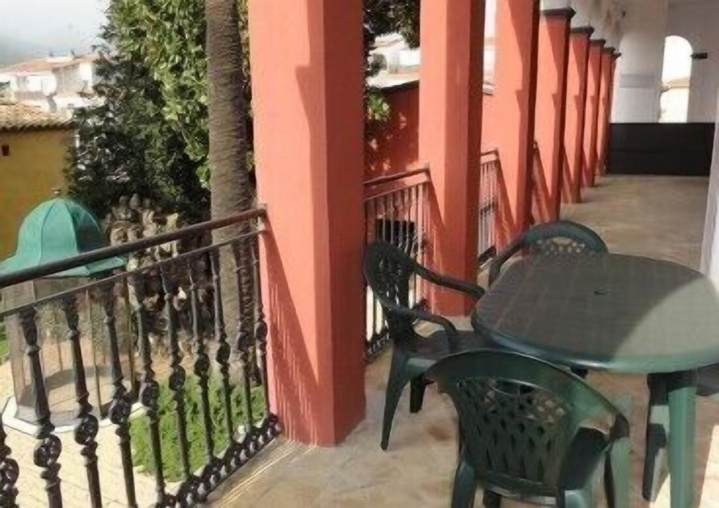 Un balcón con una mesa y sillas, barandal rojo, y vista de edificios al fondo.