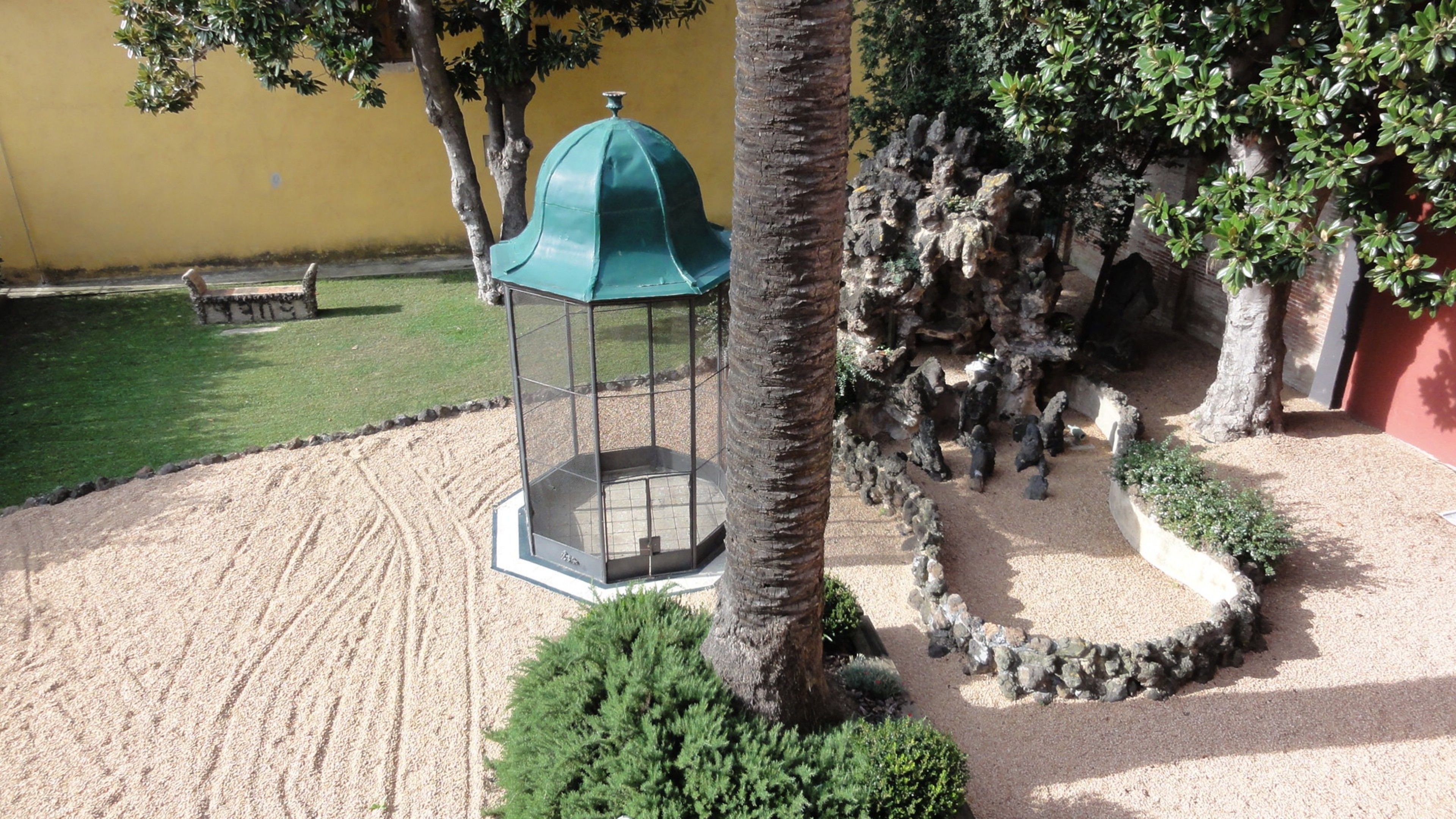 Un jardín con un pabellón, palmeras y un muro de piedra.