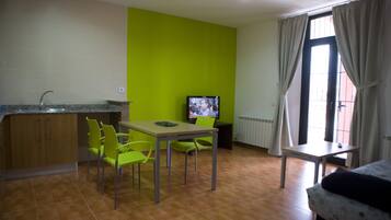 Een compacte kamer met een kitchenette, een eettafel met stoelen, een televisie en een raam met gordijnen.