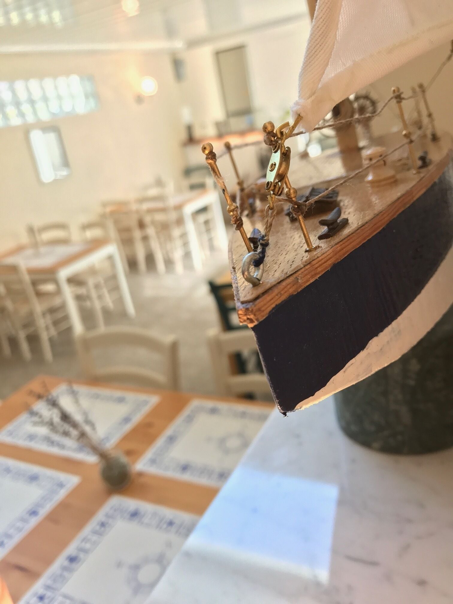 Un modelo de barco con detalles intrincados y una figura decorativa, expuesto sobre un soporte de madera en una habitación con mesas y sillas.