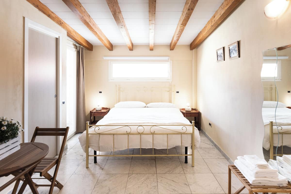Un dormitorio con cama, cabecera de madera, mesitas de noche, una pequeña mesa con una silla, una ventana y un espejo.