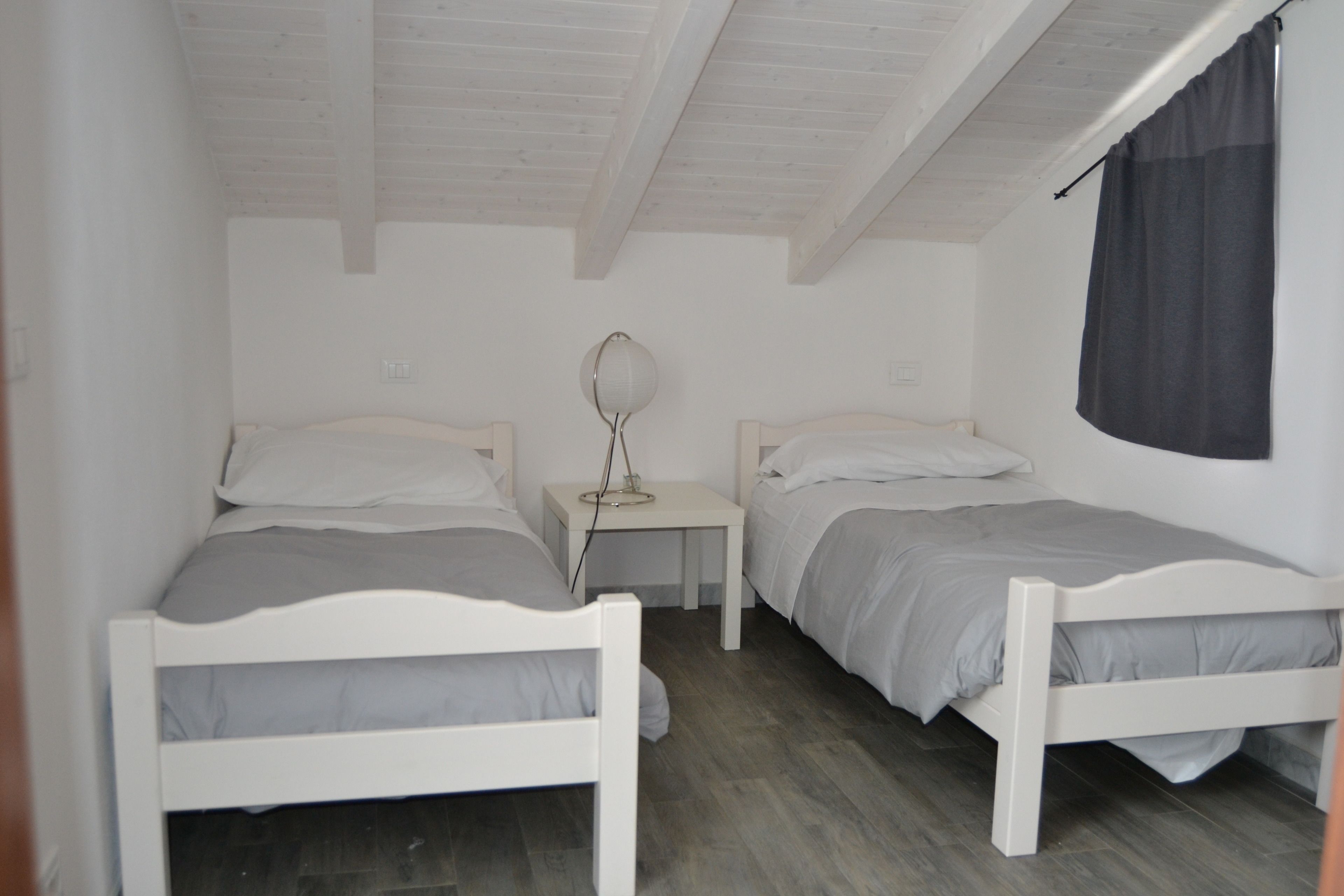 A room with two white beds, a small bedside table with a lamp, and a window with a curtain.